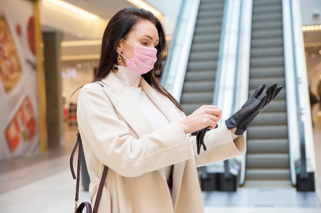 Mujer en máscara médica se pone guantes de cuero negro dentro del centro comercial o del aeropuerto