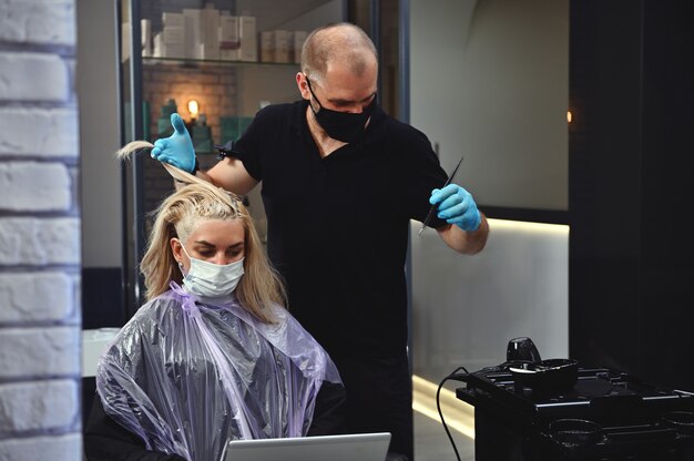 Mujer en máscara médica protectora trabaja en la computadora portátil mientras que la peluquería tiñe el mechón de cabello resaltado