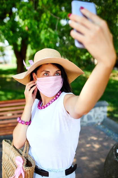 Mujer con máscara médica rosa en el parque toma selfie por teléfono móvil, la mujer con protección respiratoria está al aire libre mientras habla y tiene video chat por cámara web en el teléfono inteligente