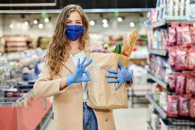 Mujer en una máscara médica sostiene una bolsa de papel con productos, verduras y firmar OK. Compras durante la pandemia de covid-19.