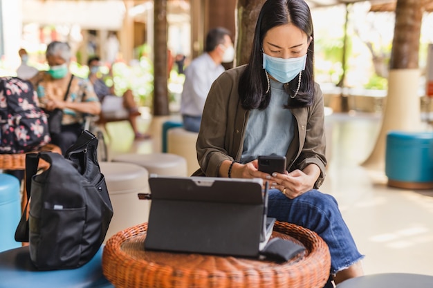 Mujer en máscara médica con teléfono celular y computadora portátil en el vestíbulo del aeropuerto.