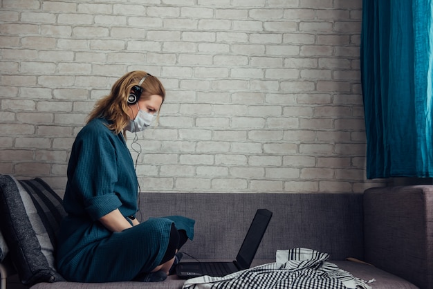 Mujer en máscara médica usando una computadora portátil en el sofá durante el trabajo remoto en casa