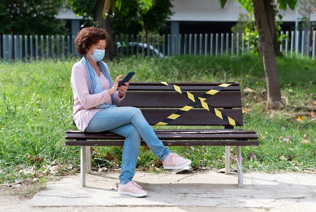 Mujer con máscara y móvil, sentada en un banco con espacio reservado para el distanciamiento social. Nueva realidad