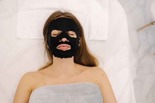 Mujer con una máscara negra hidratante de tela en la cara en el estudio de belleza cuidado facial y belleza