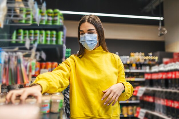 Mujer con máscara protectora y compra de alimentos en la tienda durante la epidemia del virus. Mujer joven con una máscara protectora y guantes de compras en tiempos de pandemia de virus, comprando alimentos