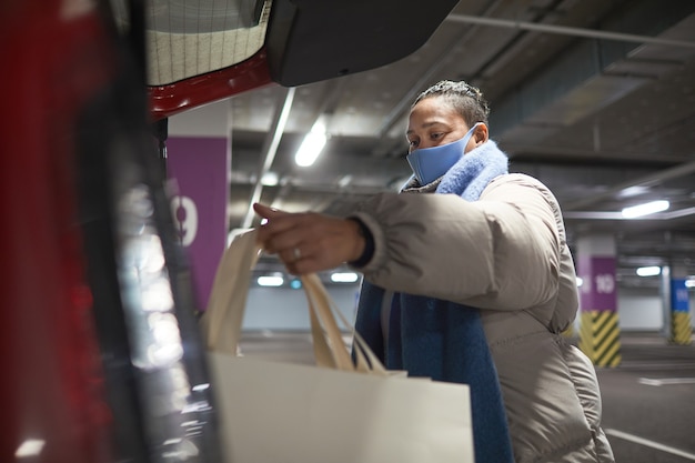 Mujer con máscara protectora empacando bolsas de compras en su automóvil mientras está de pie en el estacionamiento subterráneo