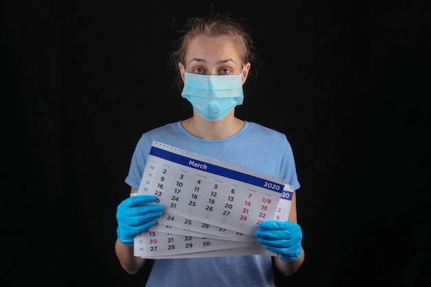 Mujer en una máscara protectora médica, guantes tienen calendario mensual en una pared negra. Cuarentena, pandemia covid-19
