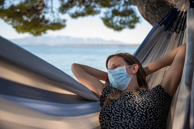 Mujer con máscara protectora médica relajándose en una hamaca junto al mar