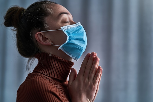 Mujer con máscara protectora con los ojos cerrados y las manos rezando, le pide a Dios sanidad y recuperación durante la enfermedad