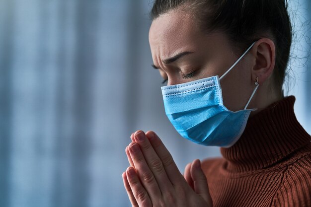 Mujer con máscara protectora con los ojos cerrados y las manos rezando, le pide a Dios sanidad y recuperación durante la enfermedad