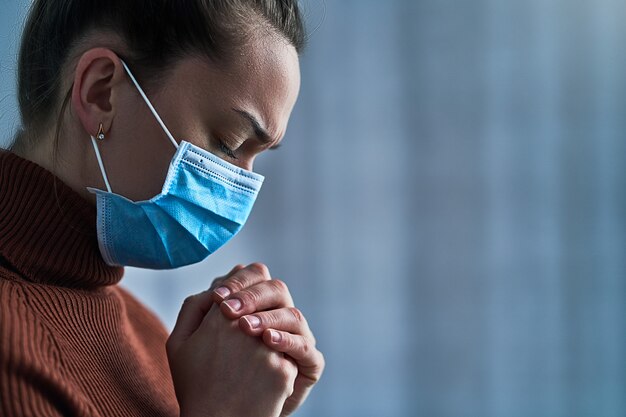 Mujer con máscara protectora con los ojos cerrados y las manos rezando, le pide a Dios sanidad y recuperación durante la enfermedad