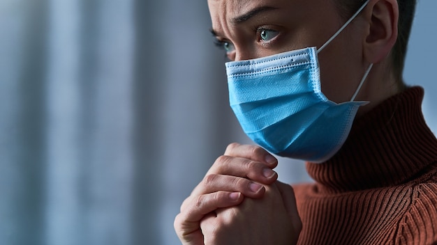 Mujer con máscara protectora con ojos tristes y manos rezando, le pide a Dios sanidad y recuperación durante la enfermedad