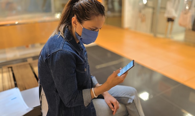 Foto mujer con máscara protectora sentada en un banco en un centro comercial y mirando el teléfono