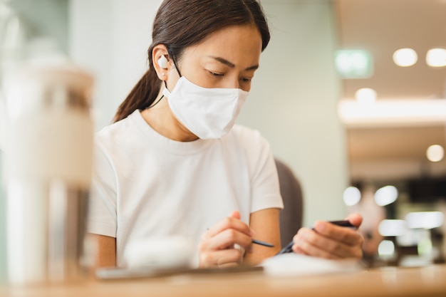 Mujer con máscara protectora trabajando en smartphone con auriculares inalámbricos en café.