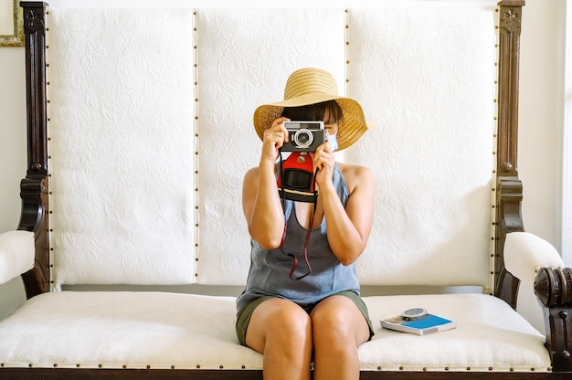 Mujer con máscara y sombrero sentado en un sillón tomando una foto y preparándose para un viaje