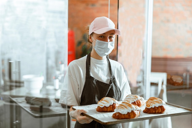 Mujer con máscara sostiene una bandeja con croissants decorados de pie en una panadería artesanal