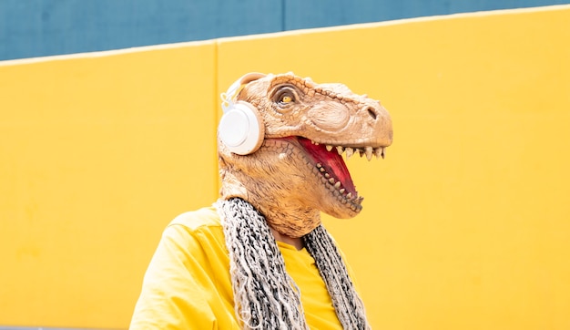 Mujer con máscara de t-rex y auriculares escucha música en una pared amarilla y azul