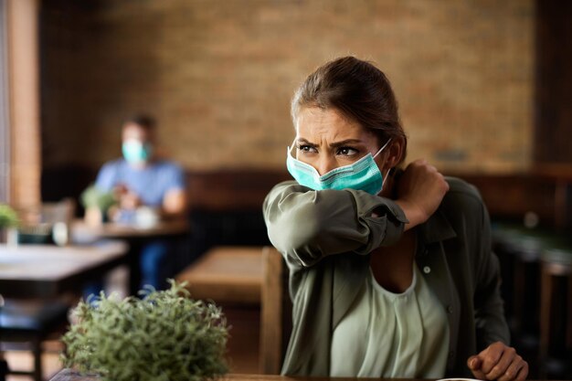 Mujer con mascarilla estornudando en el codo mientras está sentada en un café