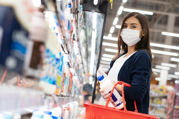 Mujer con mascarilla médica con cesta de la compra eligiendo leche diaria del estante en el supermercado