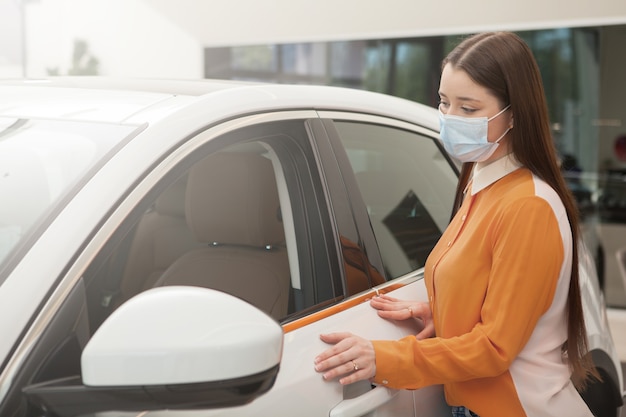 Mujer con mascarilla médica, eligiendo un auto nuevo para comprar durante la pandemia de covid-19