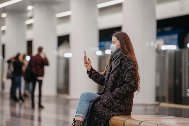 Una mujer con una mascarilla médica está sentada en el centro de la plataforma del metro con un teléfono inteligente y haciendo una selfie. Una chica con el pelo largo con una mascarilla quirúrgica mantiene distancia social en el metro.