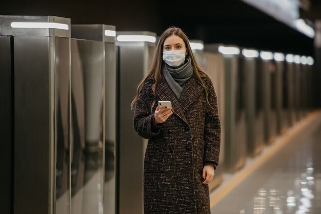 Una mujer con una mascarilla médica para evitar la propagación del coronavirus está esperando un tren y sosteniendo un teléfono celular en la estación de metro. Una niña con una máscara quirúrgica mantiene una distancia social en el metro.