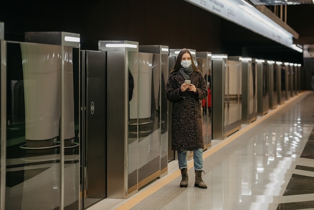 Una mujer con una mascarilla médica para evitar la propagación del coronavirus está esperando un tren y sosteniendo un teléfono en la estación de metro. Una niña con una máscara quirúrgica contra el COVID-19 mantiene la distancia social.