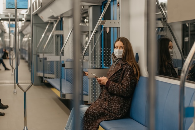 Una mujer con una mascarilla médica para evitar la propagación del coronavirus está sentada sosteniendo un teléfono inteligente en un moderno vagón de metro. Una niña con una mascarilla quirúrgica contra el COVID-19 está dando un paseo en un tren del metro.