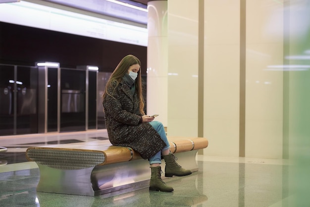 Una mujer con una mascarilla médica para evitar la propagación del coronavirus está sentada y usando un teléfono inteligente