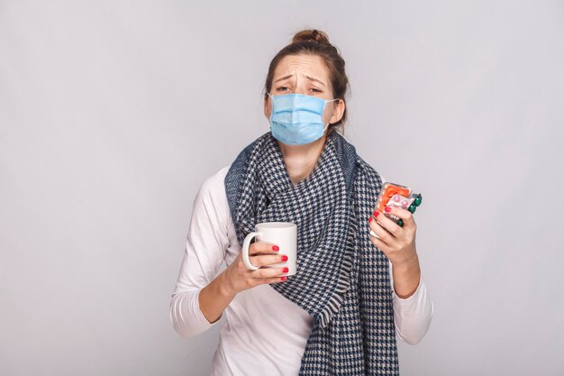 Mujer con mascarilla médica quirúrgica, llora porque estaba enferma. Sosteniendo una taza con té, muchas pastillas y antibióticos. interior, tiro del estudio, aislado sobre fondo gris