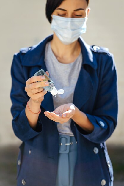 Mujer con mascarilla médica, usando un pequeño desinfectante de manos antibacteriano portátil en la calle durante el brote de Covid 19. Protección en prevención del coronavirus.