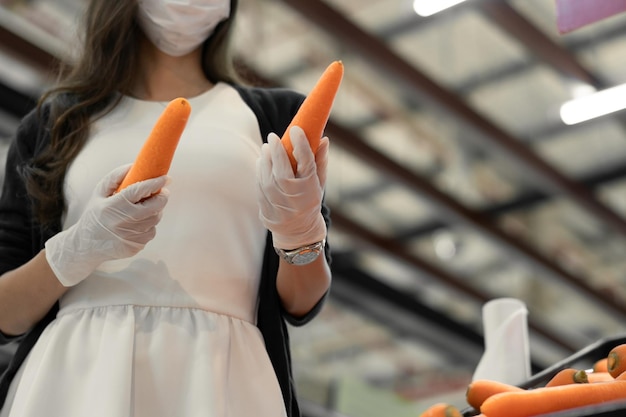 Mujer con mascarilla protectora y guante de goma sosteniendo zanahoria en el supermercado de verduras