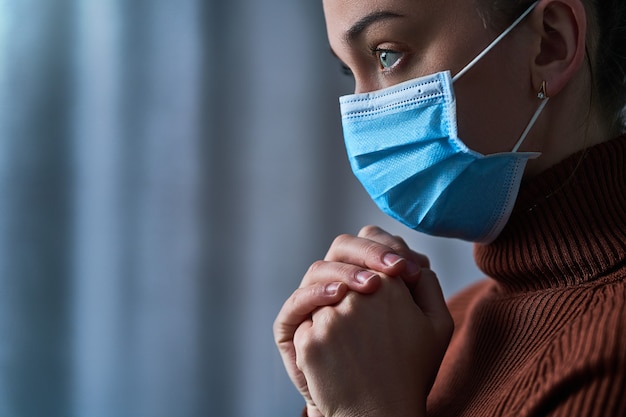 Mujer con mascarilla protectora con ojos tristes y manos rezando, le pide a Dios curación y recuperación durante la enfermedad