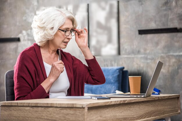 Mujer mayor ajustando anteojos mientras mira la computadora portátil