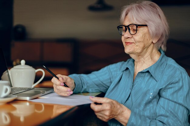 Mujer mayor con anteojos se sienta en una mesa frente a una computadora portátil Freelancer trabaja sin cambios