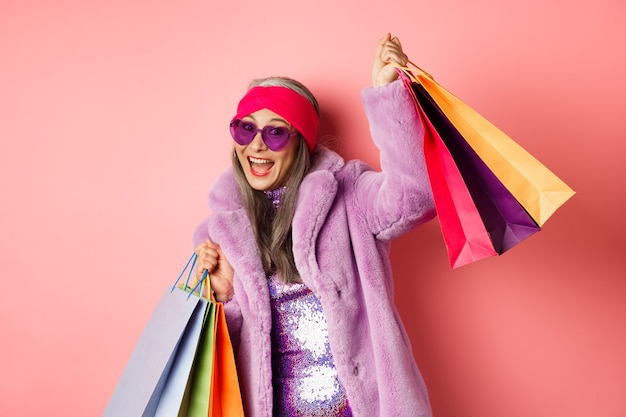 Mujer mayor asiática funky y fresca en ropa de moda bailando mientras va de compras en las rebajas, sosteniendo bolsas de papel de la tienda y divirtiéndose, fondo rosa