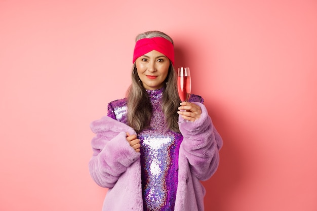 Mujer mayor asiática de moda levantando una copa de champán para felicitarlo, divirtiéndose en la fiesta y celebrando, de pie con ropa de discoteca sobre fondo rosa.