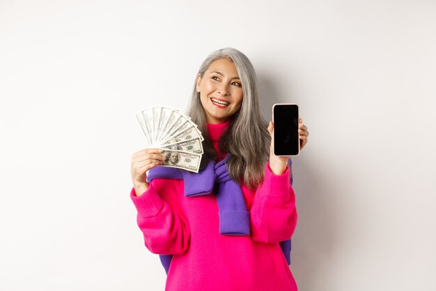 Mujer mayor asiática de moda que muestra dólares de dinero y la pantalla del teléfono inteligente en blanco, demuestra la tienda en línea, de pie sobre fondo blanco