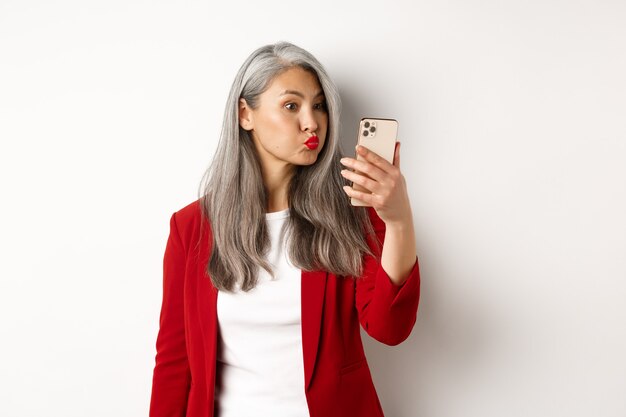 Mujer mayor asiática tonta y divertida haciendo pucheros, tomando selfie en smartphone, de pie sobre fondo blanco.