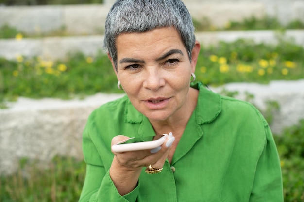 Mujer mayor atractiva que usa el teléfono móvil en el parque. Modelo femenino con cabello gris corto en ropa brillante hablando por teléfono. Ocio, actividad, concepto de tecnología.