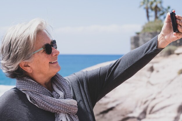 Mujer mayor atractiva en vacaciones en el mar con teléfono para selfie y sonriendo feliz jubilado relajarse