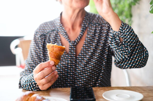 Mujer mayor bebiendo una taza de café sosteniendo un trozo de croissant sentado en un café al aire libre con un teléfono inteligente en la mesa