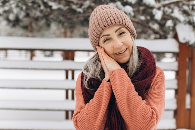 Mujer mayor con cabello gris en bufanda de suéter de punto y sombrero se ríe y calienta sus manos en invierno