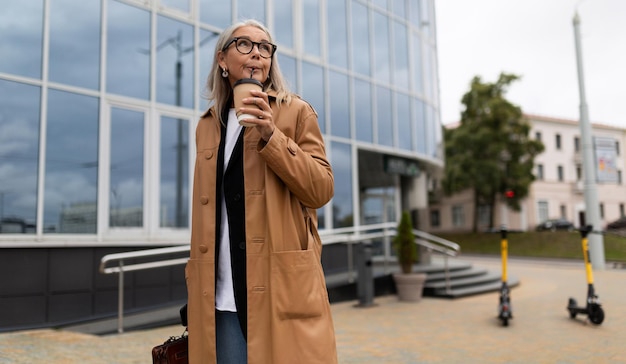 Mujer mayor camina por la ciudad con una taza de café en las manos mirando alrededor contra el