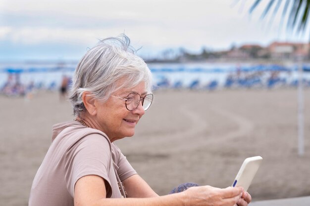 Mujer mayor caucásica sentada en la playa en un día nublado de verano usando un teléfono móvil mujer atractiva disfrutando de relajarse y vacaciones