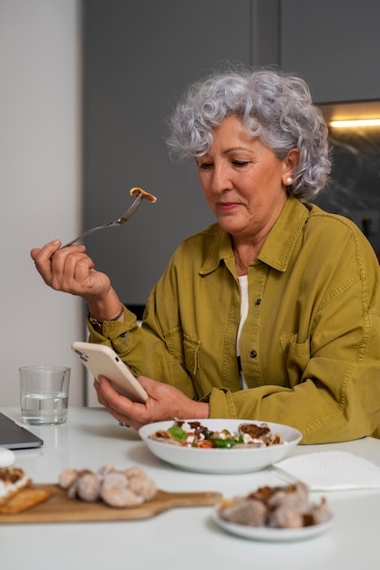 Mujer mayor comiendo plato de higos en casa y usando un teléfono inteligente