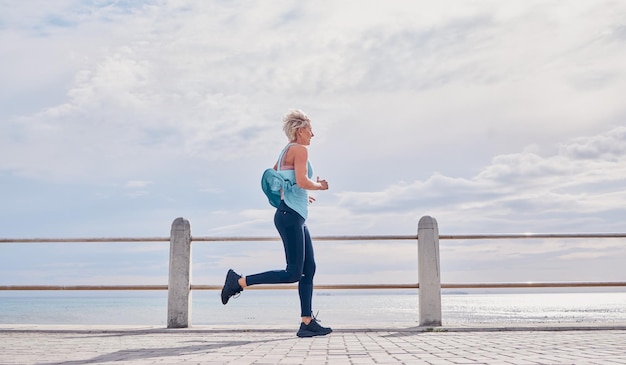Mujer mayor corriendo al aire libre en la maqueta del cielo del paseo marítimo y energía para la salud, el bienestar y el ejercicio Ejercicio femenino mayor y corredor en el océano para entrenamiento deportivo, fitness y maratón cardiovascular
