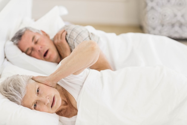 Mujer mayor despierta en la cama cubriéndose los oídos mientras su esposo ronca