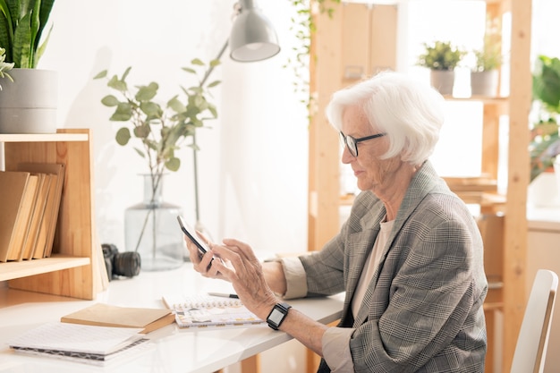 Mujer mayor con desplazamiento de dispositivo móvil o mensajería en el descanso mientras está sentado en el lugar de trabajo en la oficina