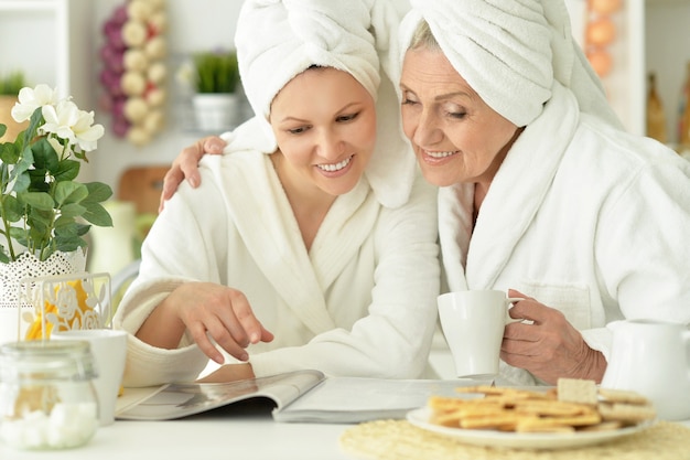 Mujer mayor e hija en una bata de baño con revista y té